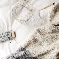 A cozy knit blanket in shades of gray and cream is spread out, accompanied by a knitting needle. Warm fairy lights are casually draped, adding a gentle glow to the scene, along with a bar of soap wrapped in a knitted cover.