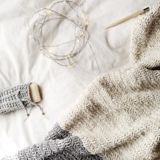 A cozy knit blanket in shades of gray and cream is spread out, accompanied by a knitting needle. Warm fairy lights are casually draped, adding a gentle glow to the scene, along with a bar of soap wrapped in a knitted cover.