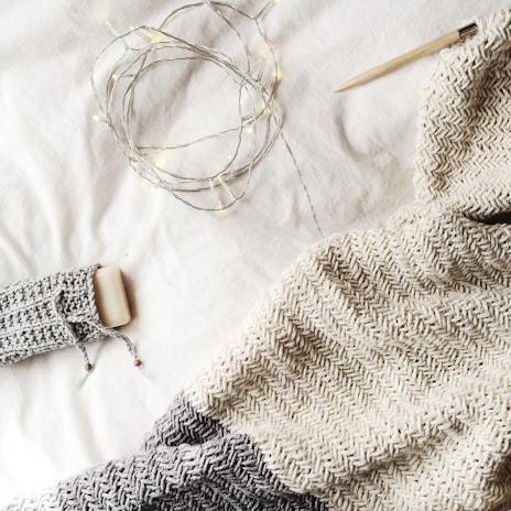 A cozy knit blanket in shades of gray and cream is spread out, accompanied by a knitting needle. Warm fairy lights are casually draped, adding a gentle glow to the scene, along with a bar of soap wrapped in a knitted cover.