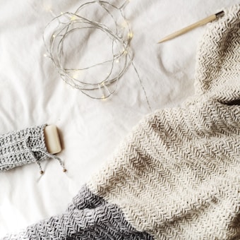 A cozy knit blanket in shades of gray and cream is spread out, accompanied by a knitting needle. Warm fairy lights are casually draped, adding a gentle glow to the scene, along with a bar of soap wrapped in a knitted cover.