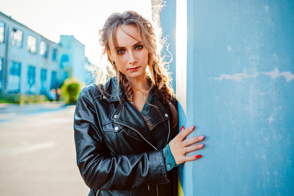 woman in black leather jacket leaning on blue wall during daytime