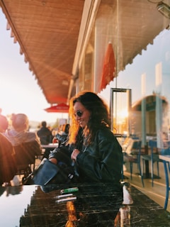 Smiling woman wearing sleek glasses, enjoying a sunny outdoor café setting.