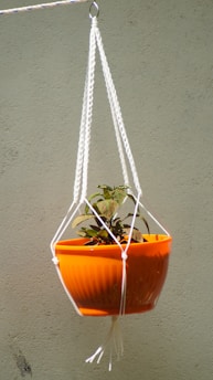 A vibrant orange planter suspended with a white macrame hanger containing a small plant with green and reddish leaves. The background is a plain beige wall.