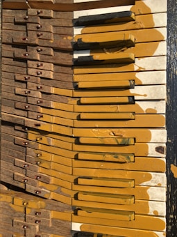 A broken piano keyboard with yellow paint splattered across the keys, some of which are stained and discolored. The wooden elements of the piano show visible wear and tear.