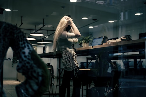 A person with long blonde hair is standing at a tall table indoors, holding their head with one hand. The space has modern decor with hanging lights and there is a laptop open on the table with greenery surrounding it. Through the glass window reflection, other people and items are faintly visible, contributing to an urban professional setting.