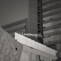 A modern urban building with large glass windows and geometric architectural design, emitting smoke from a chimney. The structure has a combination of flat and curved surfaces.