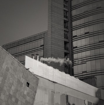 A modern urban building with large glass windows and geometric architectural design, emitting smoke from a chimney. The structure has a combination of flat and curved surfaces.
