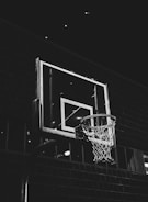 A monochrome image featuring a basketball hoop against a dark, brick wall background. The focus is on the hoop and the backboard, which is illuminated by overhead lighting, creating contrast with the surrounding darkness.