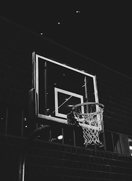 A monochrome image featuring a basketball hoop against a dark, brick wall background. The focus is on the hoop and the backboard, which is illuminated by overhead lighting, creating contrast with the surrounding darkness.