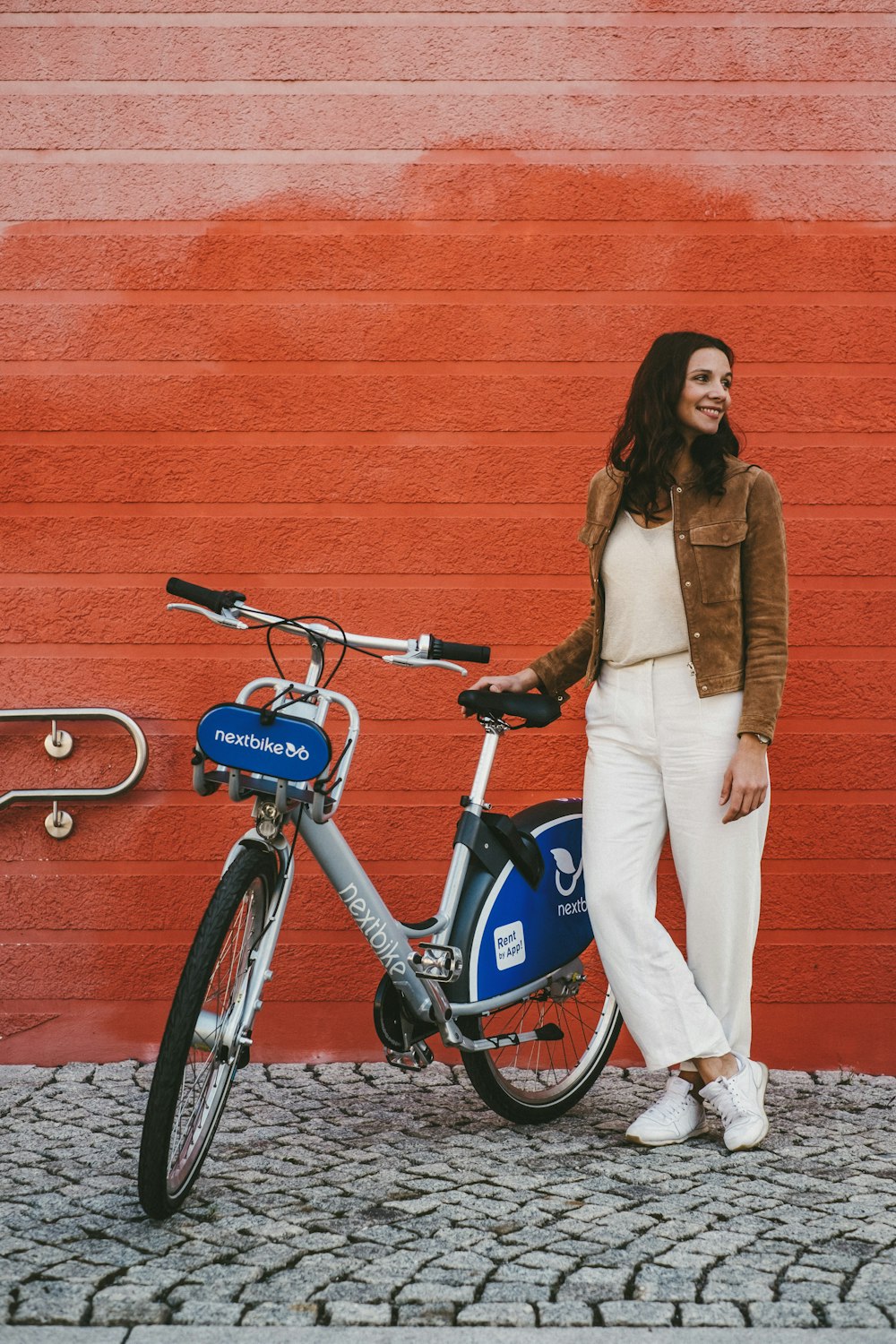 woman in brown coat and white pants standing beside blue and black bicycle