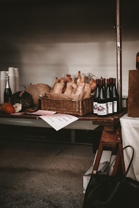 A rustic market stand displays a basket filled with multiple plucked chickens. To the right of the basket, there are bottles of red wine neatly lined up. The stand also holds pumpkins and a variety of dark and light colored produce items, alongside some disposable cups and a bottle of sparkling beverage. Labels and price tags are placed on the table, conveying an artisanal atmosphere.
