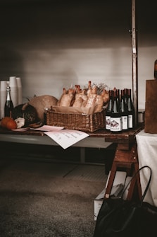 A rustic market stand displays a basket filled with multiple plucked chickens. To the right of the basket, there are bottles of red wine neatly lined up. The stand also holds pumpkins and a variety of dark and light colored produce items, alongside some disposable cups and a bottle of sparkling beverage. Labels and price tags are placed on the table, conveying an artisanal atmosphere.