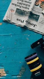 A newspaper is placed on a worn, turquoise wooden surface, with partially visible headlines in reverse. Beside the newspaper, a camera with multiple lenses stacked on top of each other is lying on the table. Parts of the old wooden surface are chipped, revealing the natural wood color beneath.