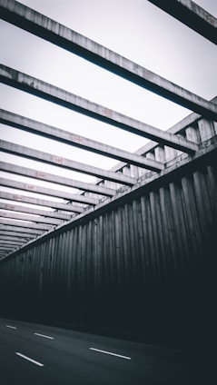 Massive concrete beams form a repetitive pattern, creating a geometric structure over a road. The design is stark and industrial, with rigid lines and a sense of depth as the structure recedes into the distance.