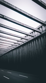 Massive concrete beams form a repetitive pattern, creating a geometric structure over a road. The design is stark and industrial, with rigid lines and a sense of depth as the structure recedes into the distance.