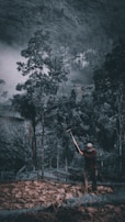 A person is working with a tool in a dense, misty forested area. Tall trees surround the individual, and the ground is rugged and uneven.
