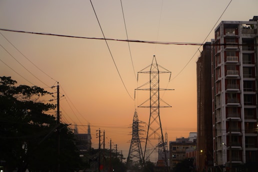 Tall electricity pylons stretching across an urban landscape, framed by buildings and trees on either side. The sky is a gradient from light to dark, suggesting either dawn or dusk. Numerous power lines crisscross the scene.