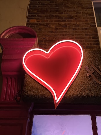 A neon red heart sign is mounted on a wall against a backdrop of dark brick and a pink architectural element. The heart is illuminated brightly, casting a warm glow on its surroundings.