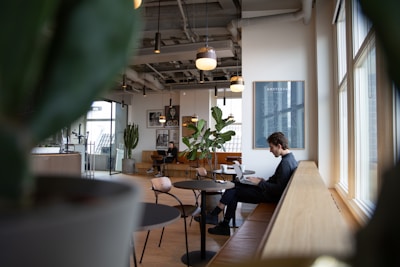 A modern, well-lit interior space featuring large windows and a seating area with wooden benches. An individual is seated on a bench working on a laptop, surrounded by stylish round tables and potted plants. The ceiling is exposed, showing industrial-style piping and hanging lights. Artwork and posters adorn the walls, adding to the contemporary design.