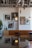 A modern interior space featuring a wall adorned with framed artworks, including abstract and portrait pieces. The seating area consists of a brown leather bench and black chairs set against a wooden table. Large plants and industrial-style lighting hang from the ceiling, paired with exposed piping that adds an urban touch to the decor.