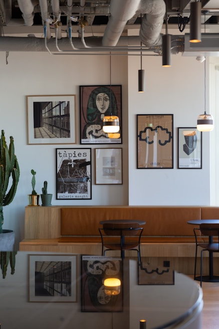A modern interior space featuring a wall adorned with framed artworks, including abstract and portrait pieces. The seating area consists of a brown leather bench and black chairs set against a wooden table. Large plants and industrial-style lighting hang from the ceiling, paired with exposed piping that adds an urban touch to the decor.
