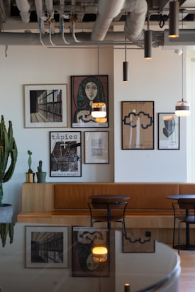 A modern interior space featuring a wall adorned with framed artworks, including abstract and portrait pieces. The seating area consists of a brown leather bench and black chairs set against a wooden table. Large plants and industrial-style lighting hang from the ceiling, paired with exposed piping that adds an urban touch to the decor.