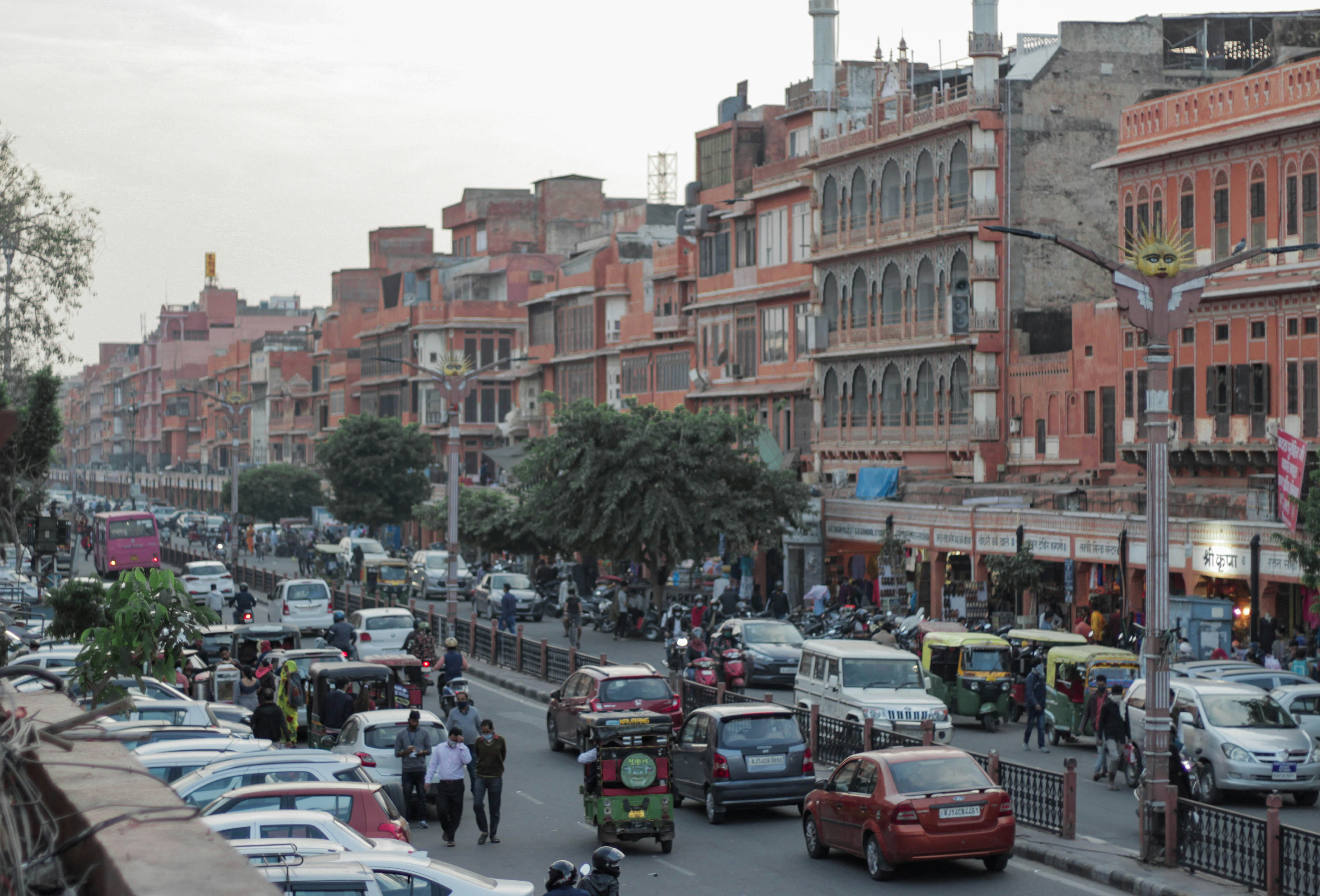 A wide shot of a Jaipur street with a large bulldozer tearing down a building, surrounded by numerous police personnel and a few onlookers. The air is dusty.