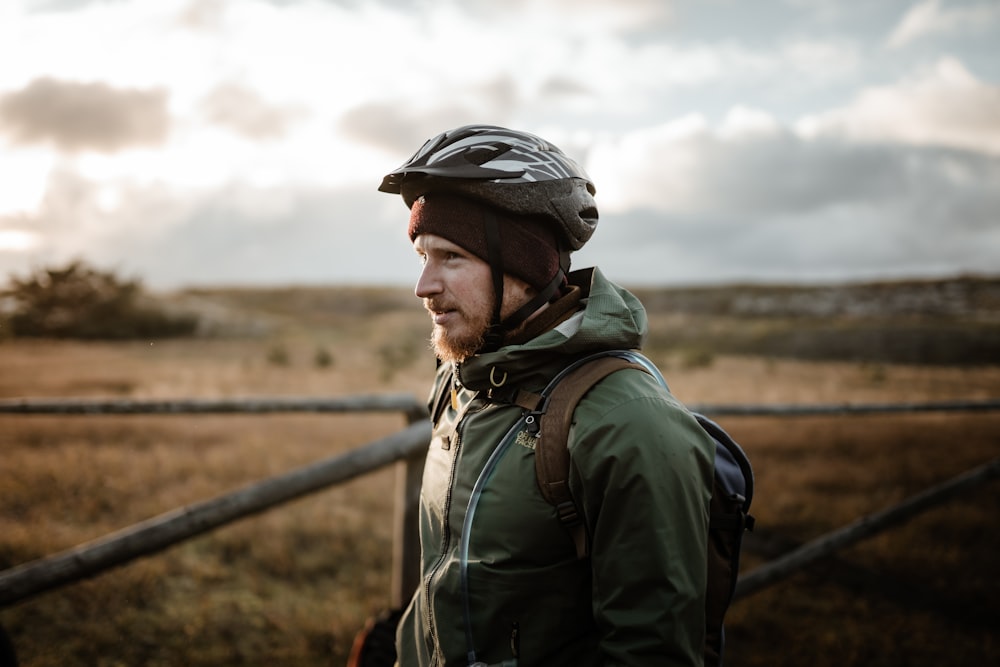 man in green jacket wearing black helmet standing near brown wooden fence during daytime