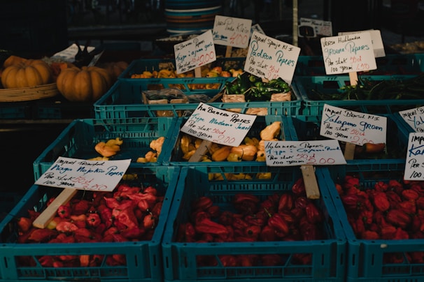 A market stall displays an assortment of colorful peppers in blue crates. Each crate is labeled with handwritten signs indicating the type and price of the peppers, such as Jalapeno, Padron, and Trinidad Naga. The peppers are green, red, and yellow, with pumpkins visible in the background.