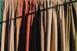 A row of hanging fabric curtains in various colors is illuminated by sunlight, casting shadows across their textured surfaces. The fabrics range from greens and browns to oranges and reds, creating a warm and earthy palette.
