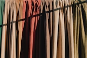 A row of hanging fabric curtains in various colors is illuminated by sunlight, casting shadows across their textured surfaces. The fabrics range from greens and browns to oranges and reds, creating a warm and earthy palette.