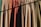 A row of hanging fabric curtains in various colors is illuminated by sunlight, casting shadows across their textured surfaces. The fabrics range from greens and browns to oranges and reds, creating a warm and earthy palette.