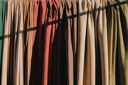 A row of hanging fabric curtains in various colors is illuminated by sunlight, casting shadows across their textured surfaces. The fabrics range from greens and browns to oranges and reds, creating a warm and earthy palette.