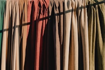 A row of hanging fabric curtains in various colors is illuminated by sunlight, casting shadows across their textured surfaces. The fabrics range from greens and browns to oranges and reds, creating a warm and earthy palette.
