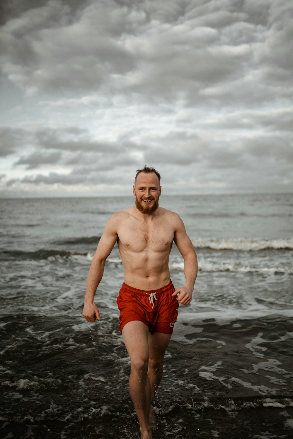 topless man in blue shorts standing on beach shore under cloudy sky during daytime