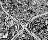 An aerial view of an urban landscape featuring a complex network of highways intersecting with several overpasses. Surrounding the highways, there are densely packed residential areas and commercial buildings. The scene captures a detailed layout of roads and infrastructure in a black and white color scheme.