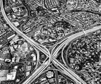 An aerial view of an urban landscape featuring a complex network of highways intersecting with several overpasses. Surrounding the highways, there are densely packed residential areas and commercial buildings. The scene captures a detailed layout of roads and infrastructure in a black and white color scheme.