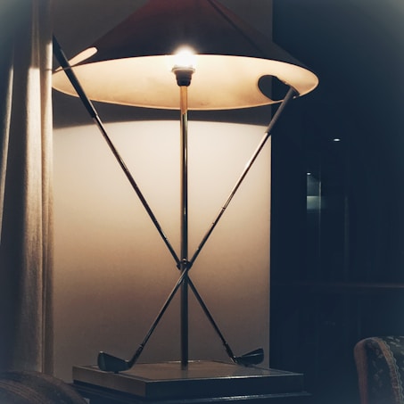 A lamp with a minimalist design, featuring a conical shade and geometric metal framework. The base rests on a small table with decorative elements around. A soft glow emanates from the lamp, casting subtle shadows on the wall and creating a cozy ambiance.