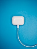 A white wireless earphones charging case is plugged in with a cable against a bright blue background. The minimalist setup highlights the contrast between the white case and the vibrant background.