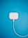 A white wireless earphones charging case is plugged in with a cable against a bright blue background. The minimalist setup highlights the contrast between the white case and the vibrant background.