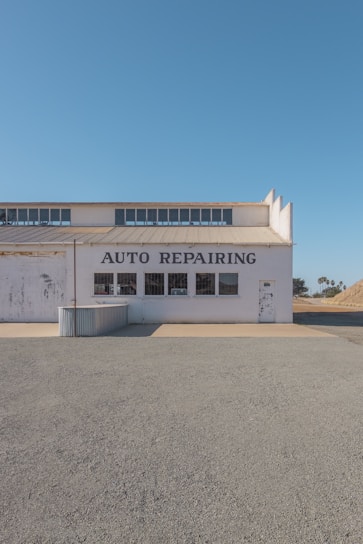 A single-story building with the words 'AUTO REPAIRING' displayed prominently above a series of windows. The structure appears weathered, with some paint peeling off, indicating age and exposure to the elements. The scene is set on a clear day with a bright blue sky, and the ground in front of the building is covered in gravel or dirt. A small, corrugated metal structure is attached to the side of the building.