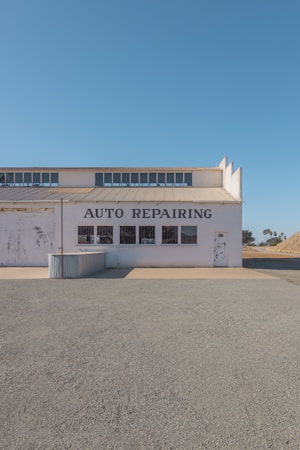 A single-story building with the words 'AUTO REPAIRING' displayed prominently above a series of windows. The structure appears weathered, with some paint peeling off, indicating age and exposure to the elements. The scene is set on a clear day with a bright blue sky, and the ground in front of the building is covered in gravel or dirt. A small, corrugated metal structure is attached to the side of the building.