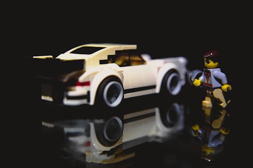 A LEGO model of a white sports car is placed on a reflective surface, creating a mirrored effect. Next to it stands a LEGO figure dressed in casual attire, appearing to hold a tool. The scene is set against a dark background, emphasizing the vivid colors of the LEGO pieces.