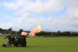 A group of people is gathered around a military cannon positioned on a grassy field. The cannon is firing, with a bright flash and smoke visible. Vehicles and trees are seen in the background under a partly cloudy sky.