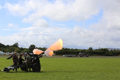 A group of people is gathered around a military cannon positioned on a grassy field. The cannon is firing, with a bright flash and smoke visible. Vehicles and trees are seen in the background under a partly cloudy sky.