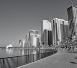 A waterfront cityscape featuring a row of modern high-rise buildings and a clear sky. Palm trees line the pathway beside the water, and a white building with minarets is visible in the distance. Construction cranes are also present, indicating ongoing development.