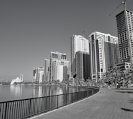 A waterfront cityscape featuring a row of modern high-rise buildings and a clear sky. Palm trees line the pathway beside the water, and a white building with minarets is visible in the distance. Construction cranes are also present, indicating ongoing development.