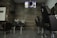 A modern hair salon interior with empty black styling chairs arranged in a row. A wall-mounted television displays a scene above two hair-washing stations. The salon features dark wooden wall panels and gray tiled flooring.