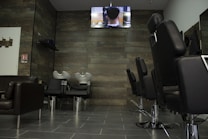 A modern hair salon interior with empty black styling chairs arranged in a row. A wall-mounted television displays a scene above two hair-washing stations. The salon features dark wooden wall panels and gray tiled flooring.