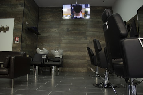 A modern hair salon interior with empty black styling chairs arranged in a row. A wall-mounted television displays a scene above two hair-washing stations. The salon features dark wooden wall panels and gray tiled flooring.